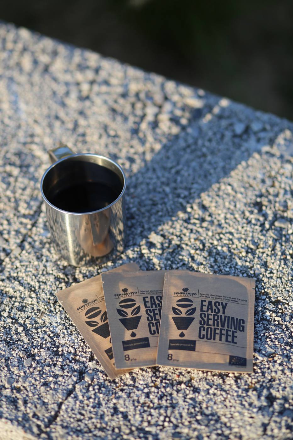 Three Semprevivo Bio Series drip sachets fanned beside a steel mug of black coffee on a sunlit stone ledge