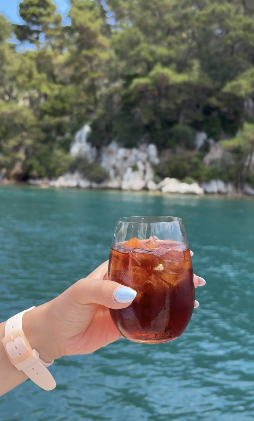 Iced coffee in a glass held up against turquoise water and green cliffs
