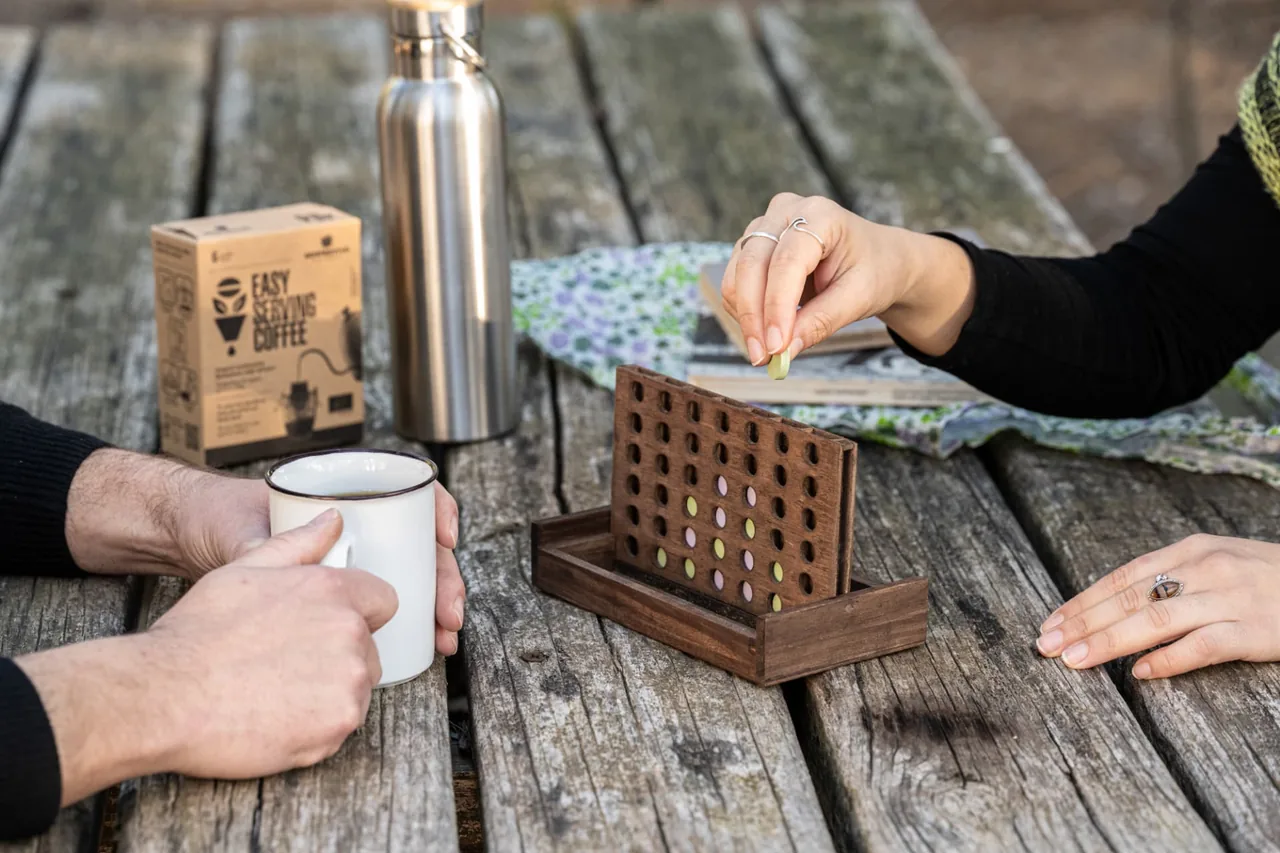 Two people playing a wooden board game at a picnic table with a mug of drip coffee and the Semprevivo box