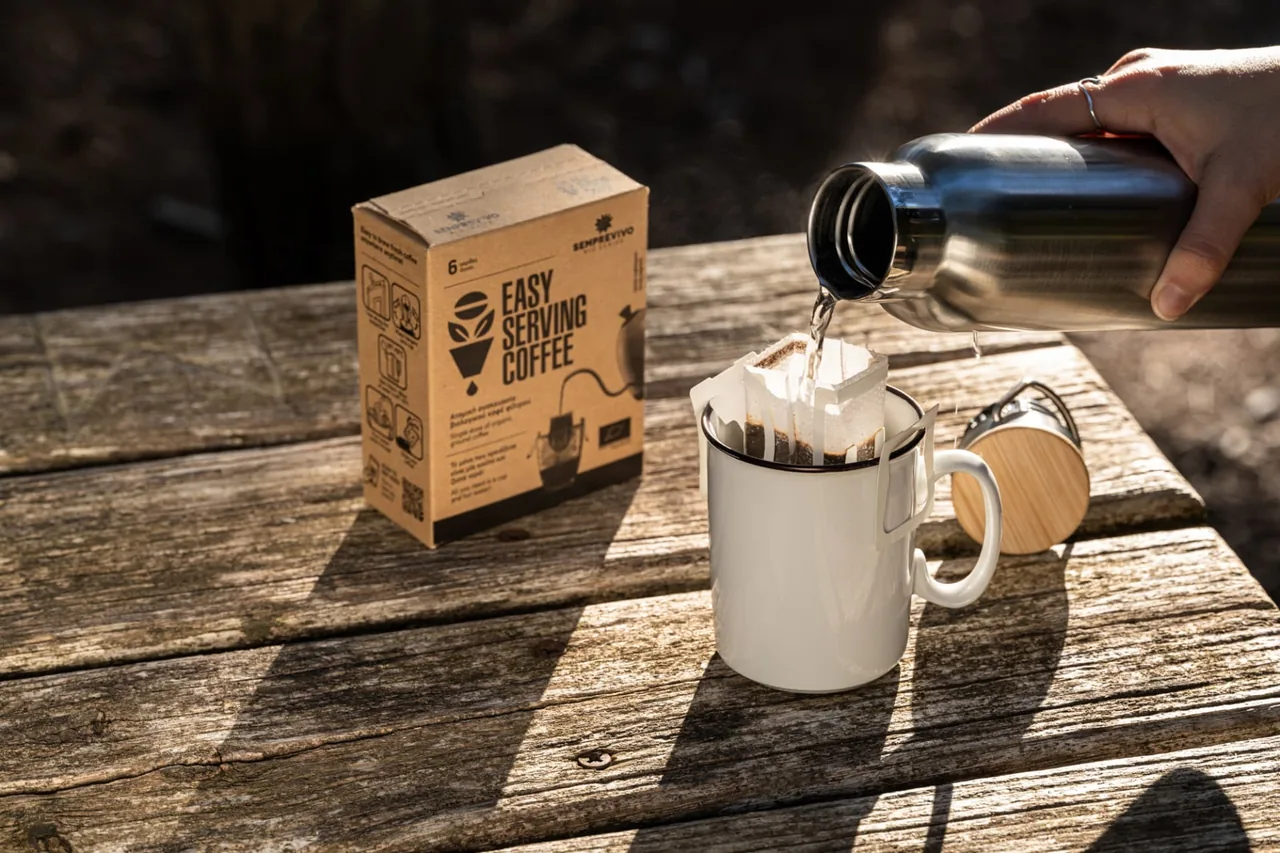 Hot water poured from a steel flask over a Semprevivo drip bag in an enamel mug on a wooden picnic table