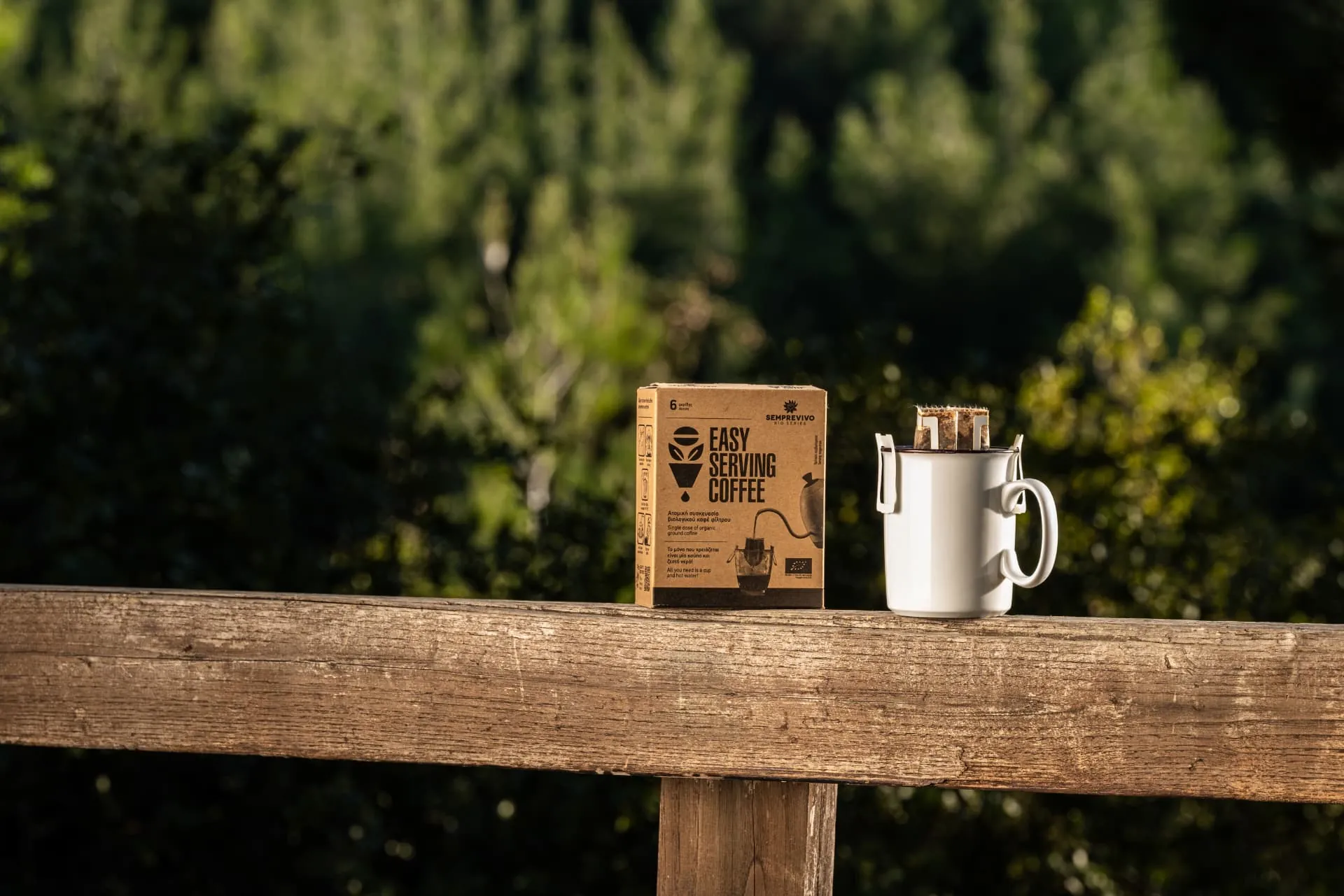 Semprevivo Easy Serving Coffee box beside a white mug with a drip bag, on a wooden fence in the forest