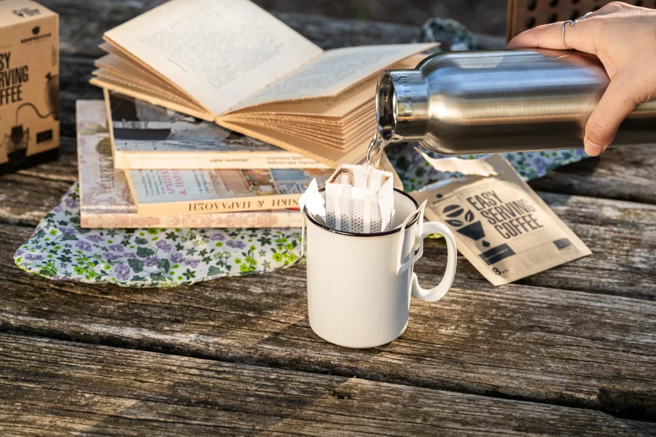Pouring hot water from a flask into a drip bag in an enamel mug beside a stack of books